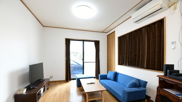 A living room with a flat-screen TV on a wooden stand, a blue sofa, a wooden coffee table, and a large window with curtains.