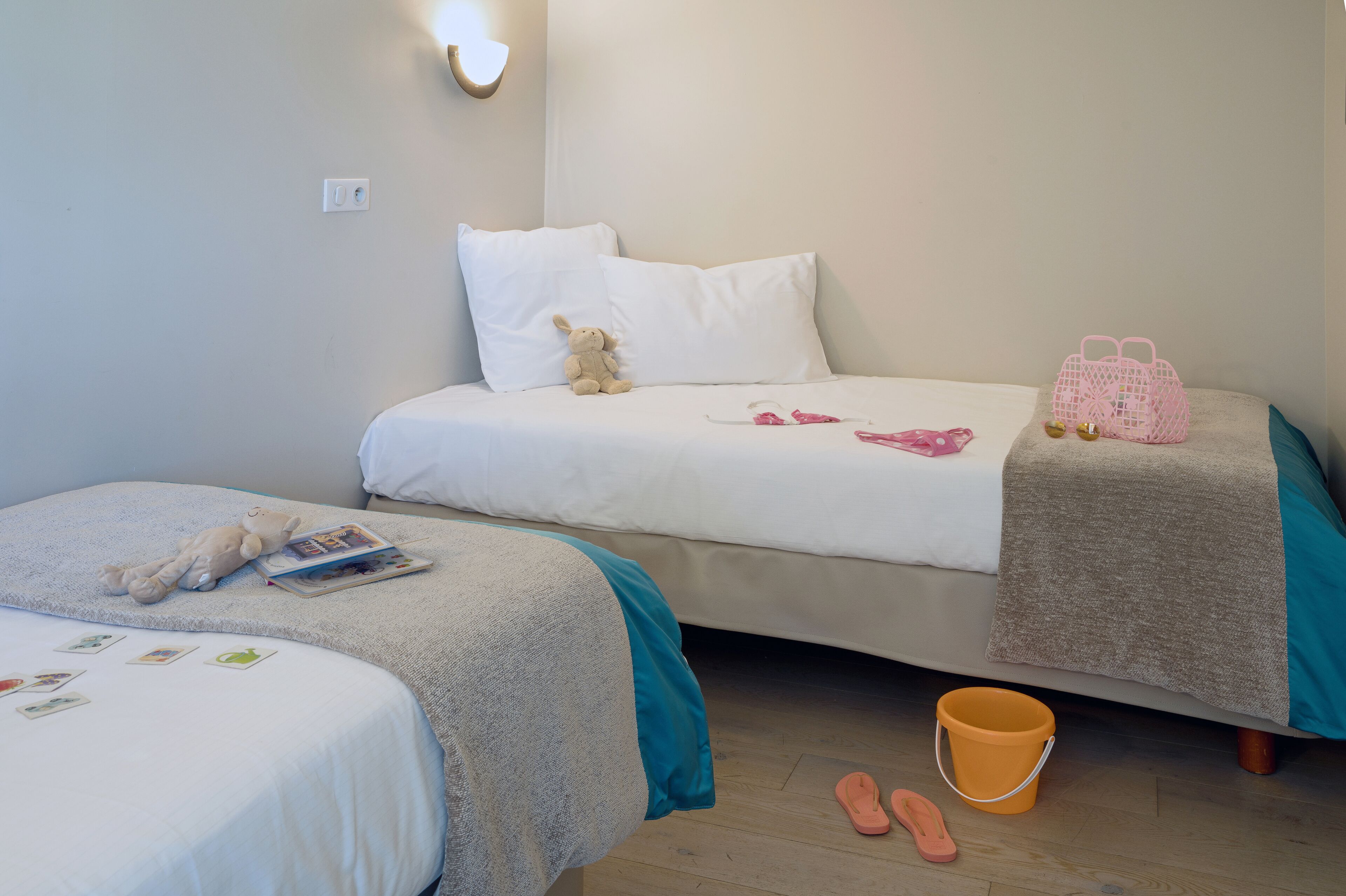 A bedroom with two beds, a teddy bear, a pink bag, a bucket, and slippers.