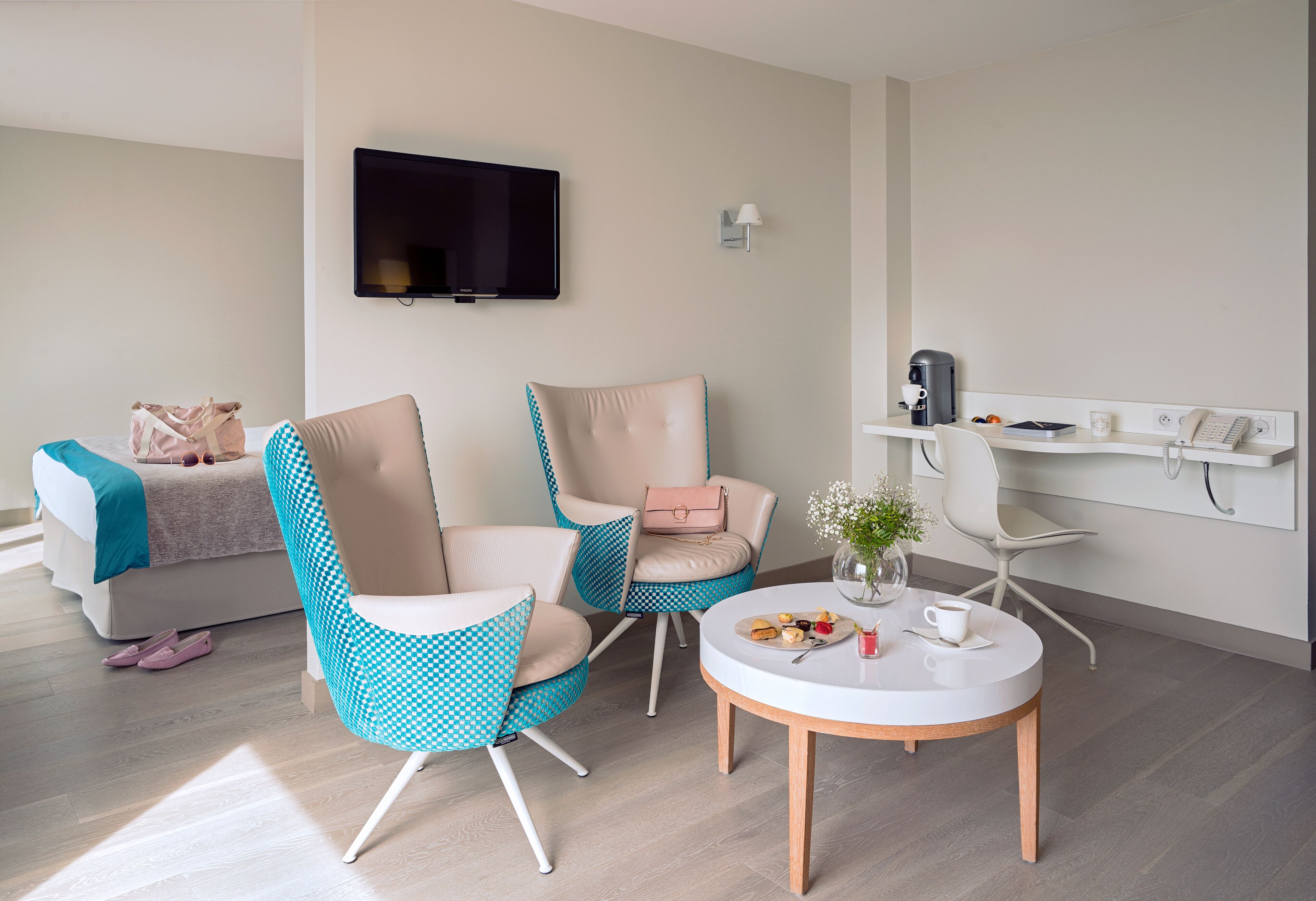 A modern living room with a sofa, two patterned chairs, a round coffee table, a TV mounted on the wall, a desk with a phone and a coffee machine, and a vase with flowers.