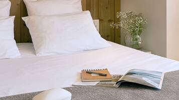 A neatly made bed with white linens, a bedside lamp, a small vase with flowers, and a pair of sunglasses on a grey pillow.