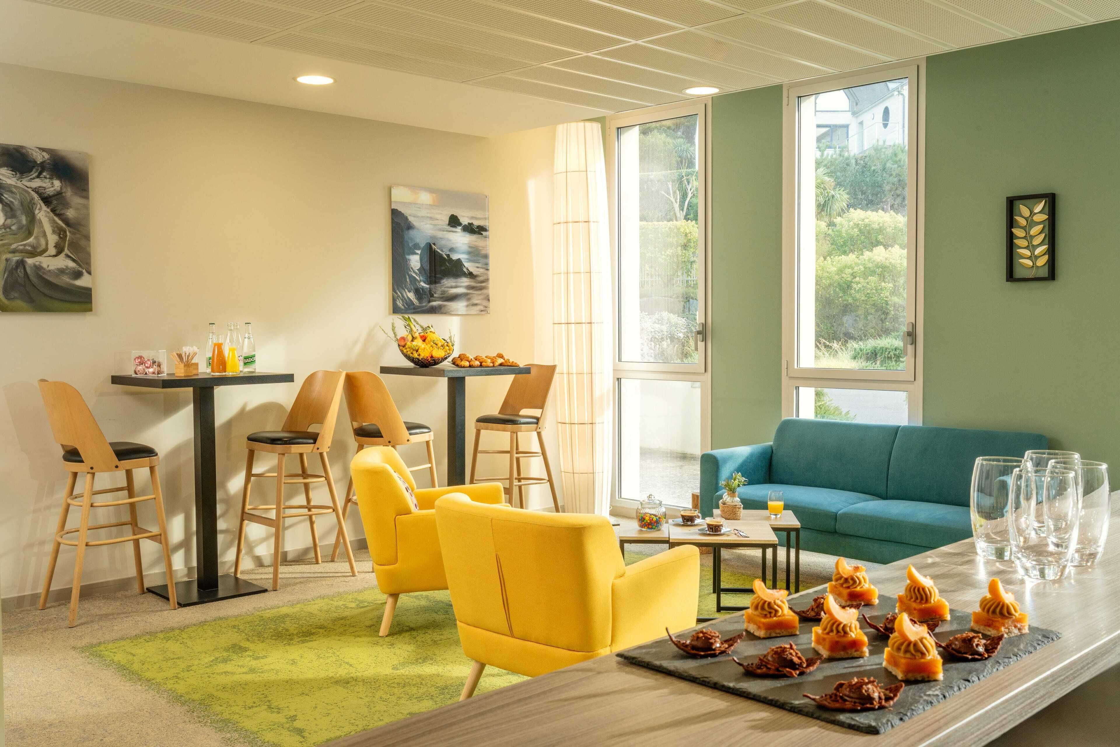 Una habitación de hotel moderna con comedor, sillas amarillas, un sofá azul y una mesa con comida y bebidas.