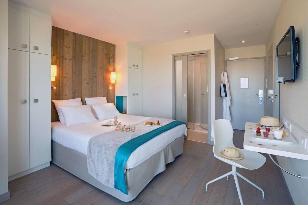 Une chambre d’hôtel moderne équipée d’un lit, d’un bureau avec une chaise, d’un miroir et d’un espace douche.
