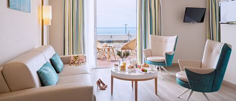A modern hotel room with a beige sofa, teal patterned armchair, and a round coffee table with breakfast items. A TV is mounted on the wall, and the room has a view of the beach through large windows.