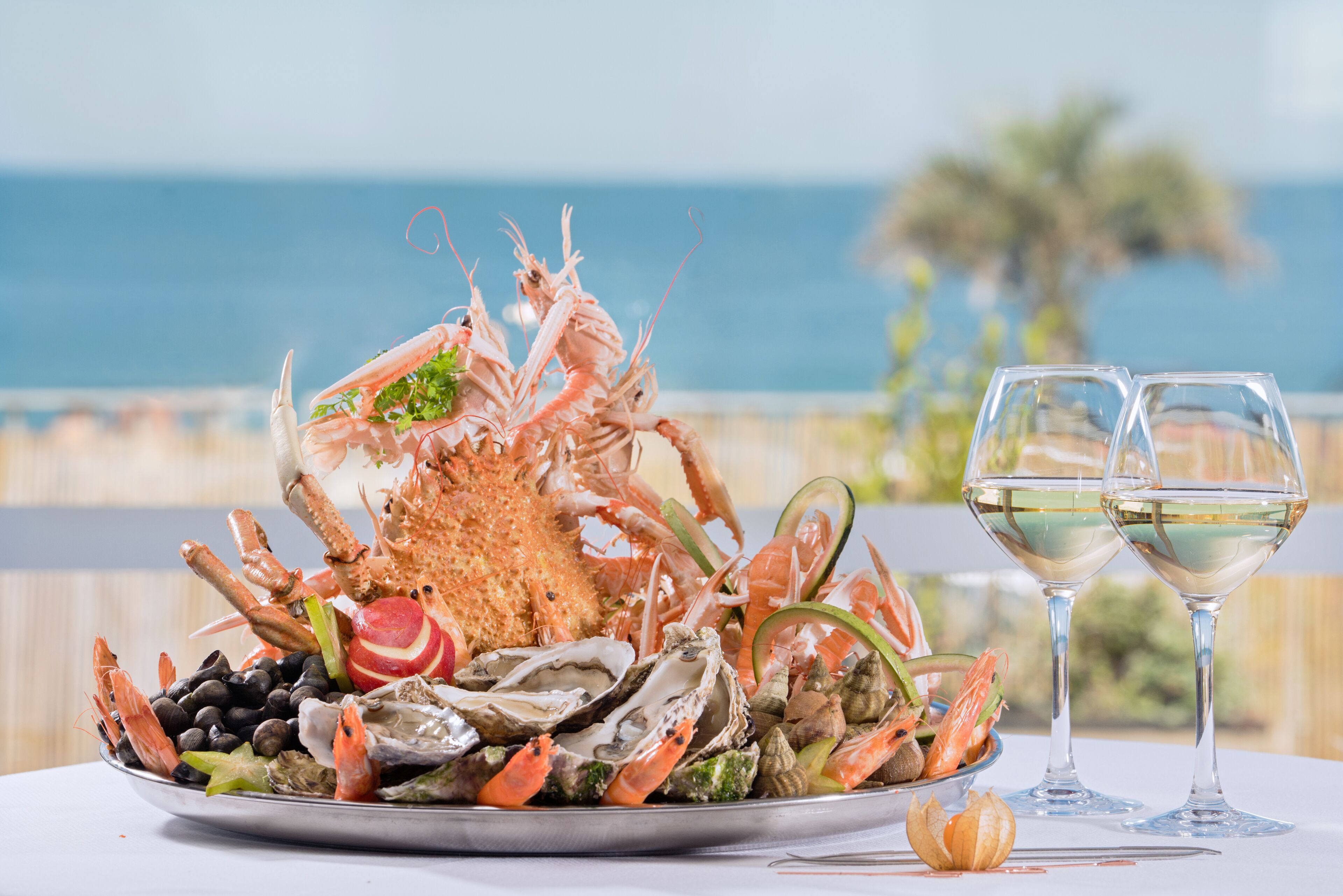 Un plateau de fruits de mer comprenant des huîtres, des crevettes et d’autres fruits de mer, accompagné de deux verres de vin blanc, le tout servi dans un cadre offrant une vue imprenable sur l’océan.