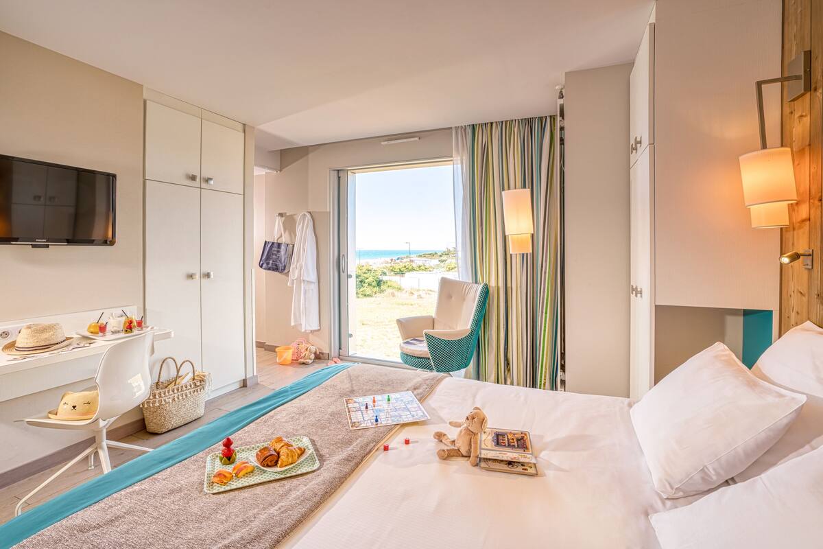 Une chambre d’hôtel avec un lit, un bureau avec une télévision, une chaise, une table avec de la nourriture et une vue sur la plage.
