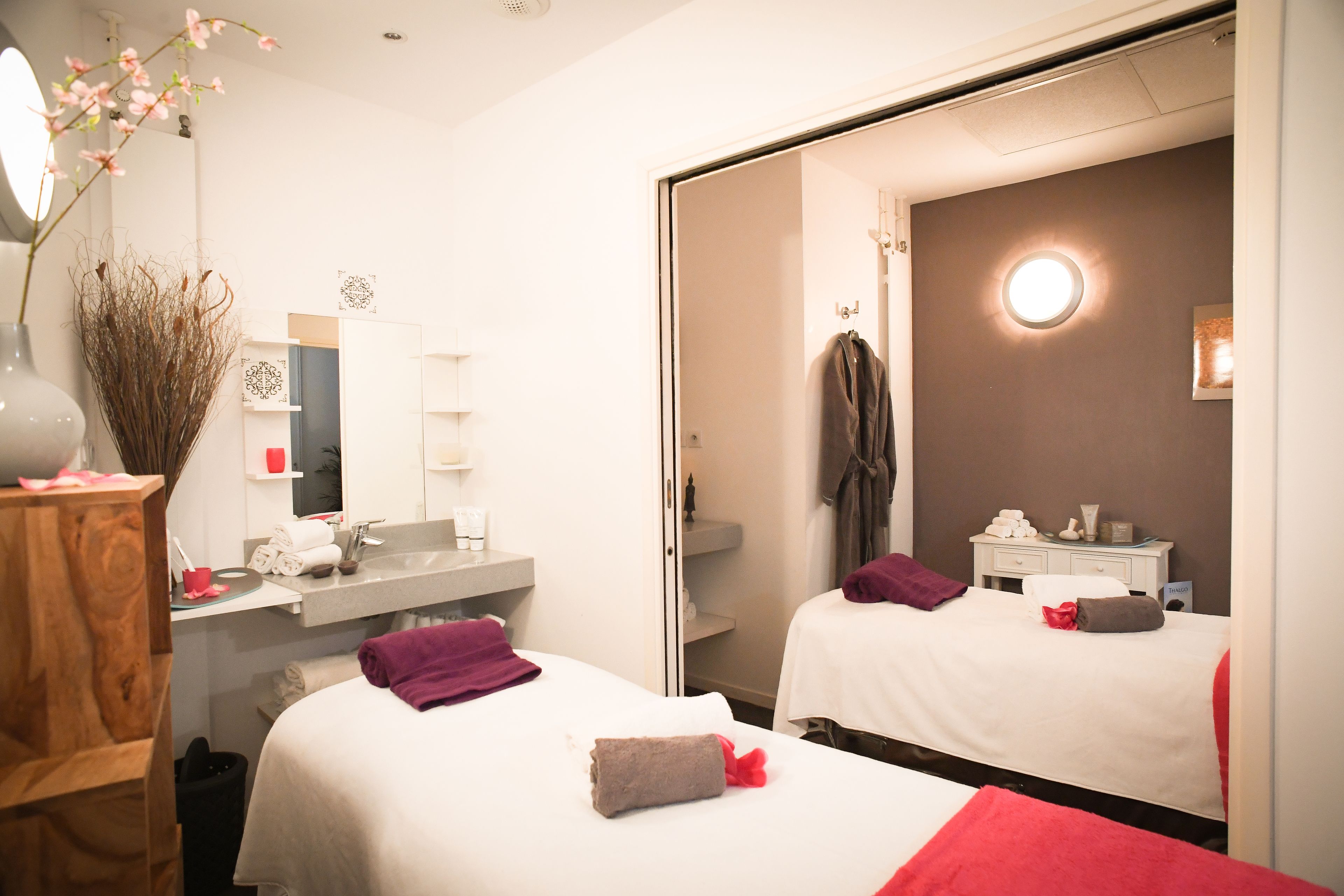 A hotel room with two beds, a bathroom with a mirror, and a bedside table with a vase and a lamp.