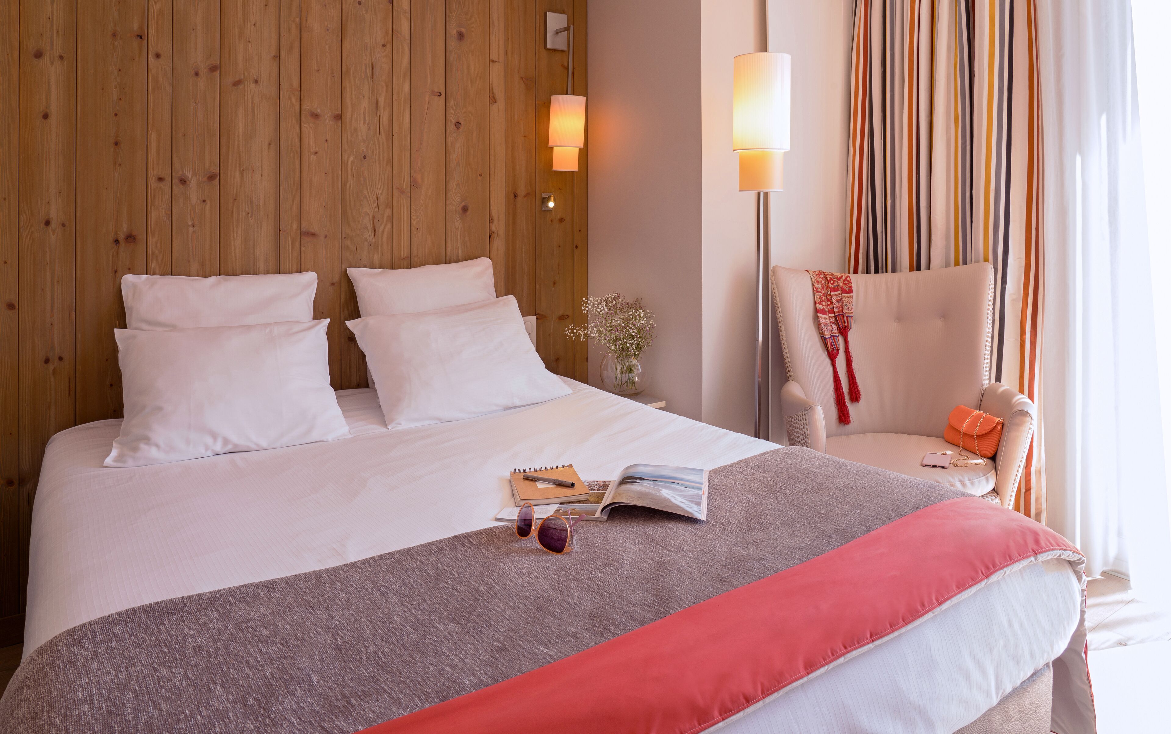 Un lit bien fait, avec du linge de lit blanc, un plaid rouge, un livre et des lunettes de soleil posés sur le lit. On y voit une tête de lit en bois, une table de chevet avec une lampe et une chaise recouverte d’un plaid rouge.