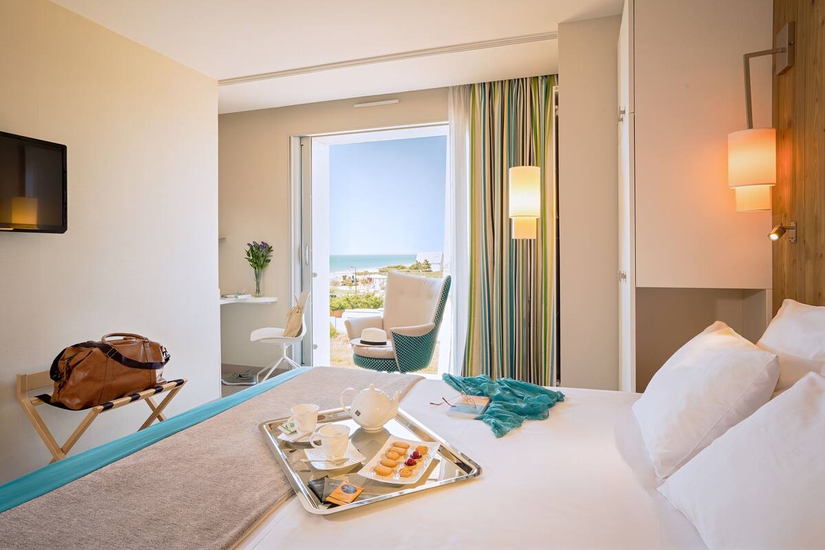 Une chambre d’hôtel avec un lit, un plateau contenant des articles pour le petit-déjeuner, une chaise, une vue sur l’océan et un balcon avec des rideaux.