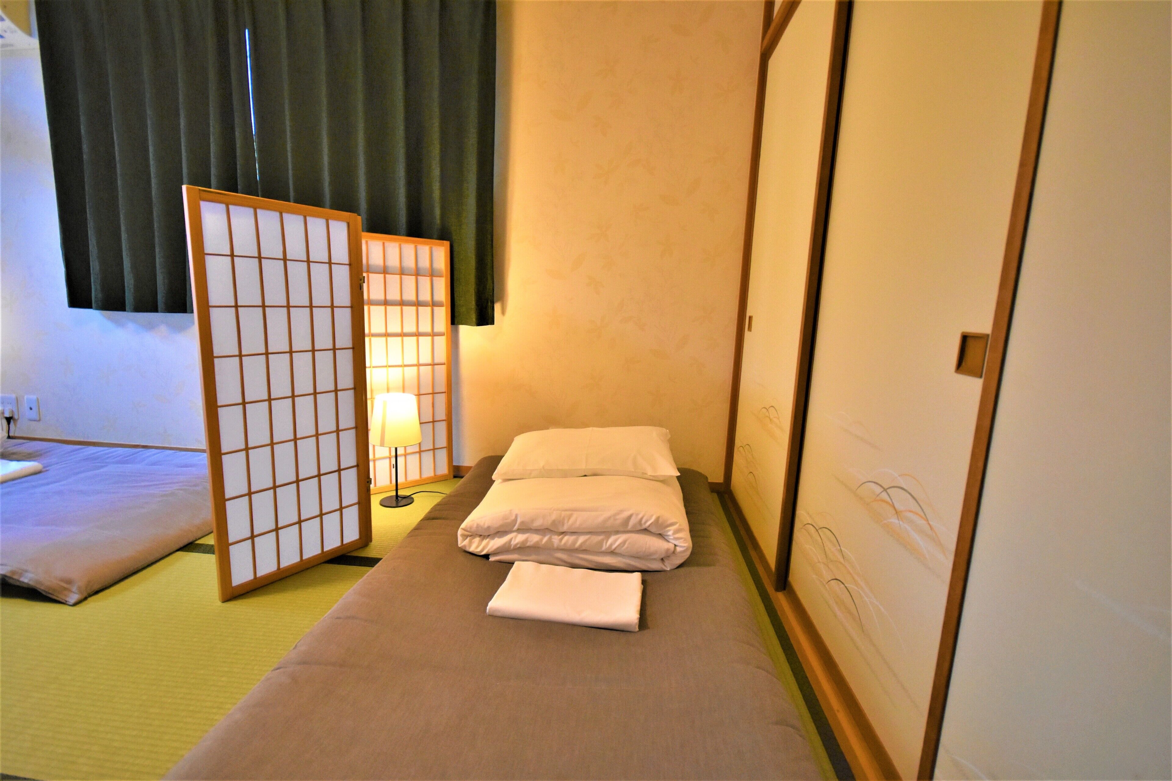 A traditional Japanese room with tatami flooring, a low bed with white bedding, a shoji screen, and a wall with a subtle floral pattern.