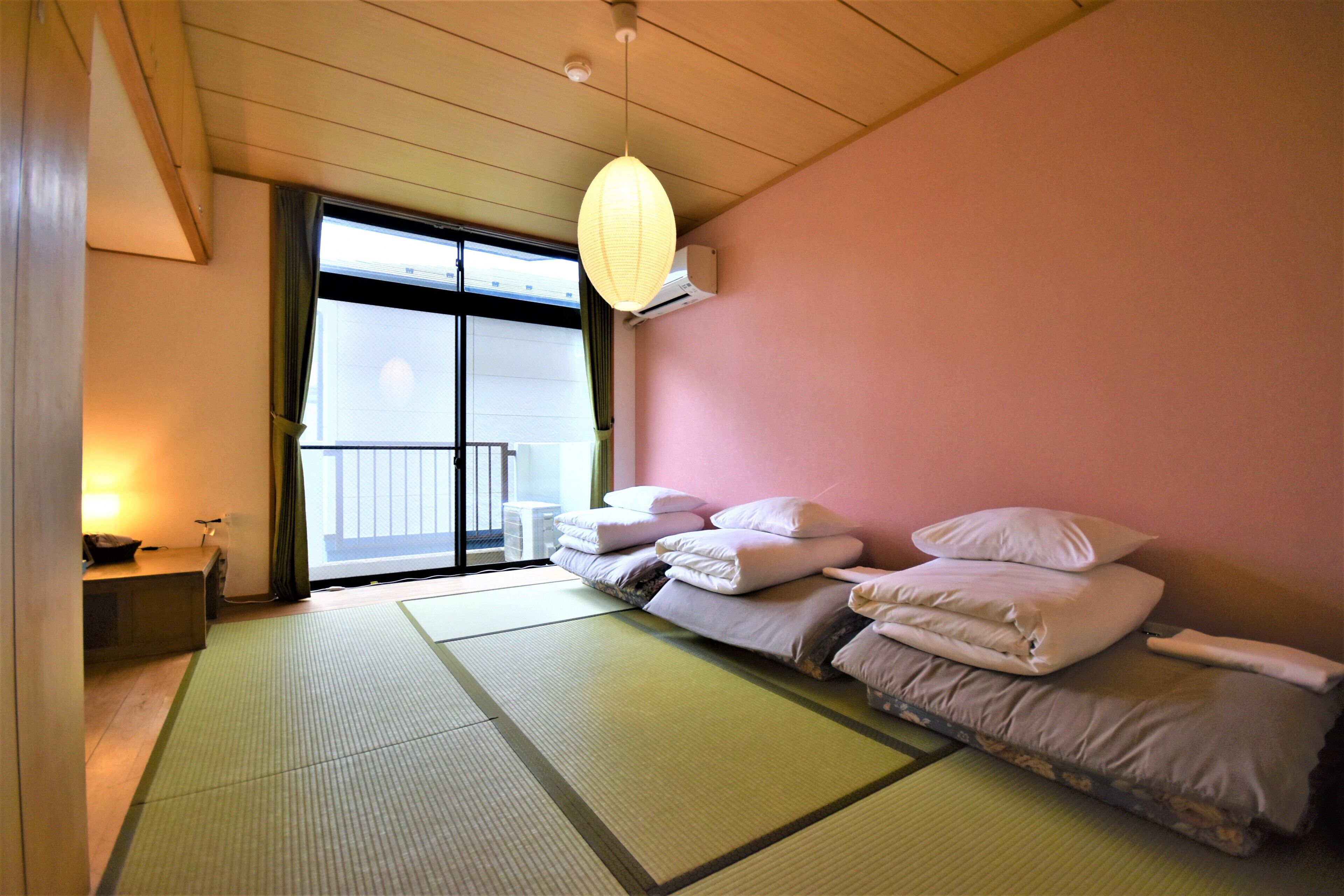 Quarto com tatames, futon com travesseiros, porta de vidro deslizante para uma varanda e um lanterna de papel suspensa.