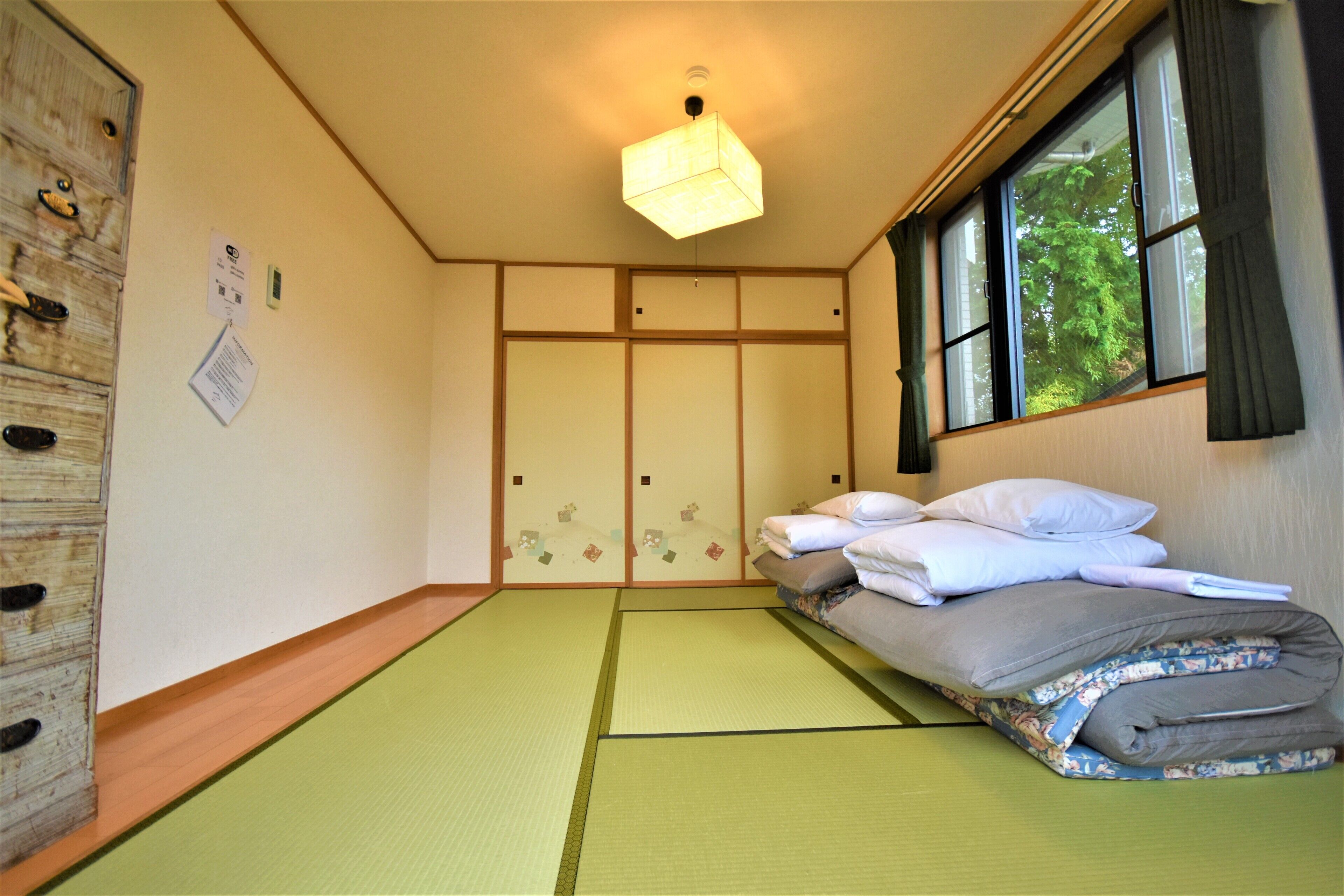 A room with tatami flooring, a bed with pillows, a hanging light fixture, and a window with curtains.