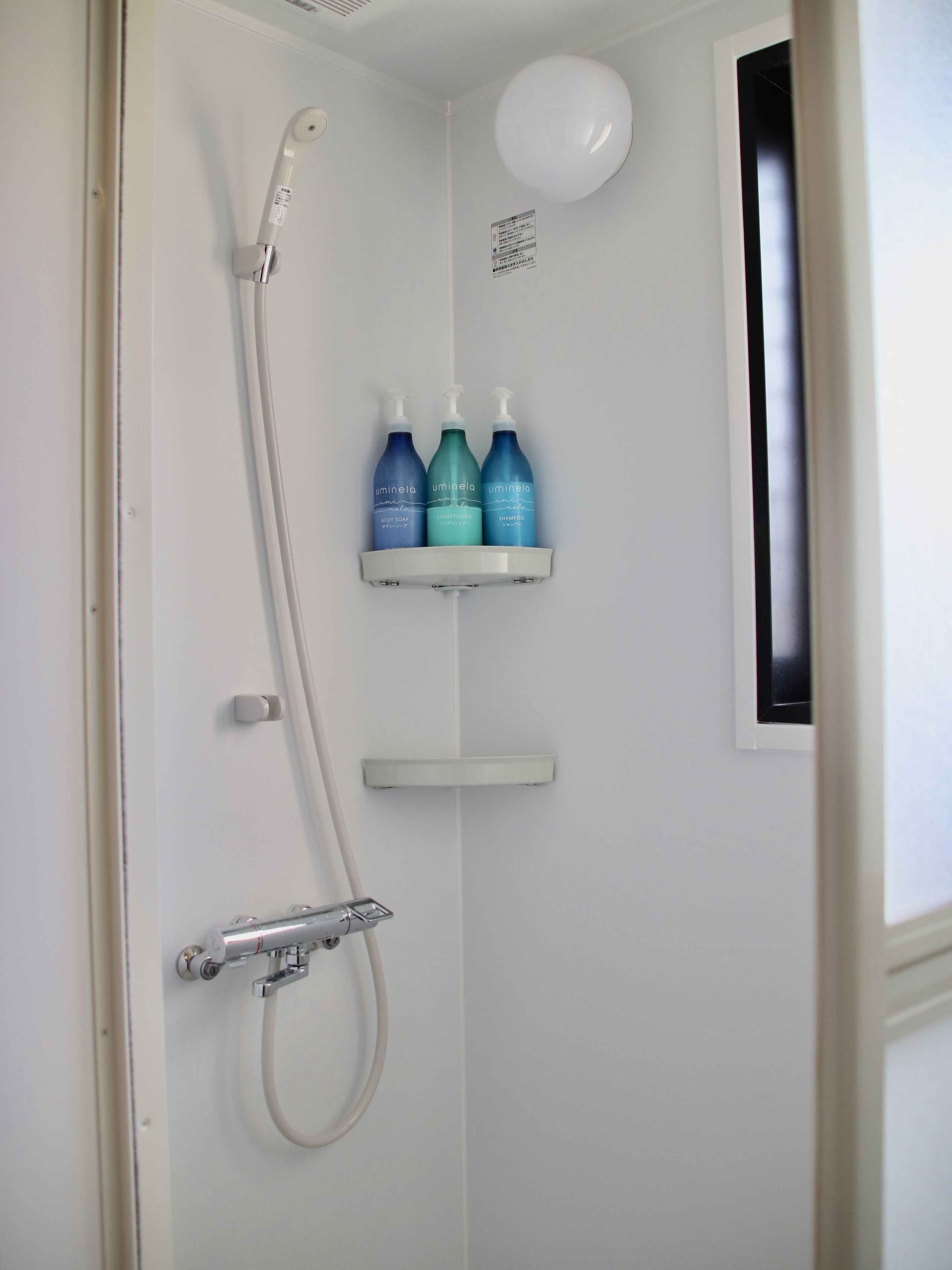 Zona de ducha con cabezal de ducha manual, grifo de pared y tres botellas de productos 'uminela' en una repisa.