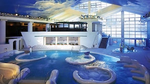 An indoor swimming pool with multiple water features, a central staircase, and a large glass wall providing a view of the sky.