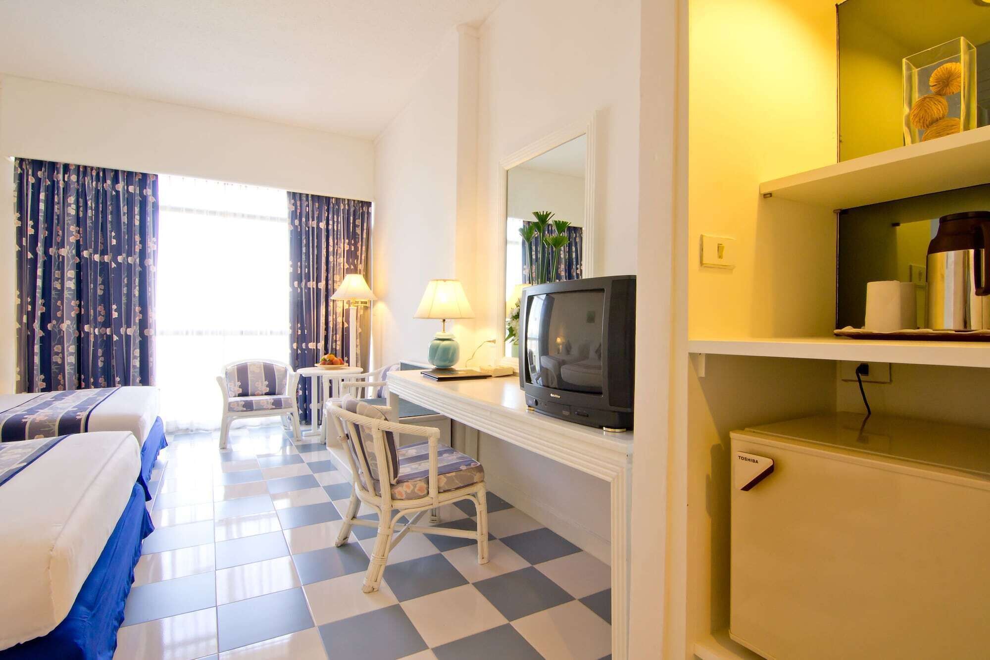 A hotel room with a checkered floor, a bed, a desk with a television, a chair, and a lamp.