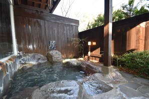 A hot spring area with stone steps leading into a clear pool, surrounded by wooden structures and greenery.