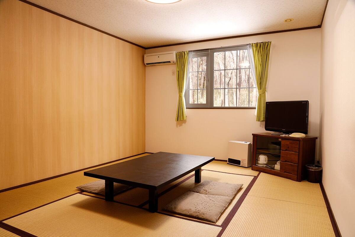 Una habitación con suelo de tatami, una mesa baja, un televisor sobre un soporte de madera y una ventana con cortinas.