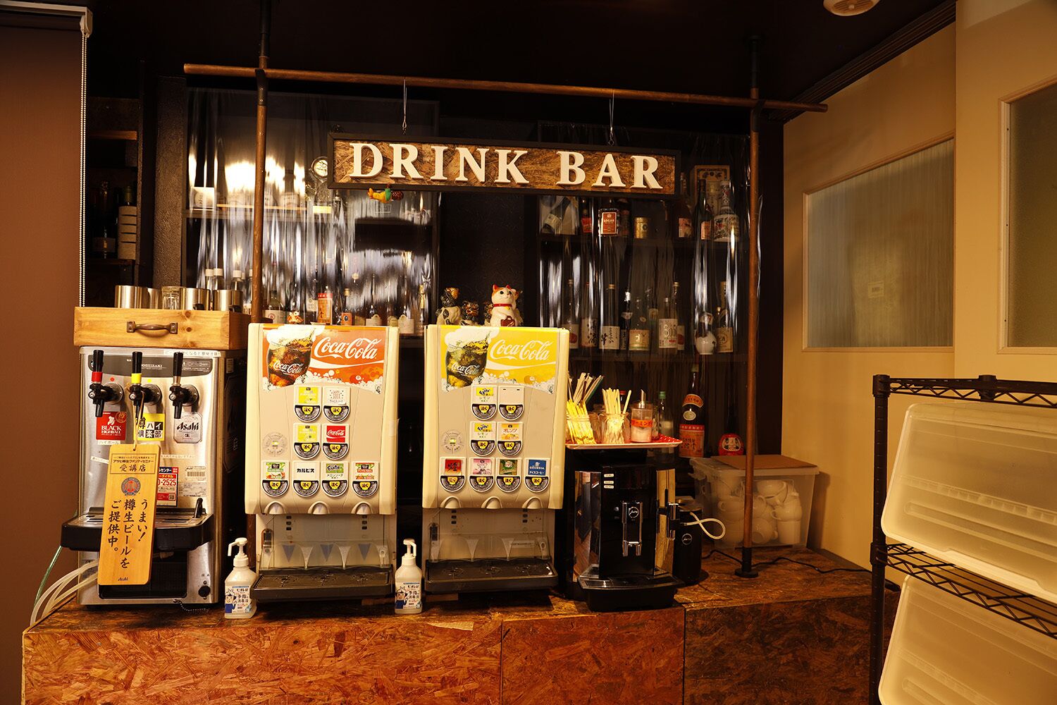 A drink bar with two Coca-Cola vending machines, a coffee machine, and a variety of bottles and containers on shelves.