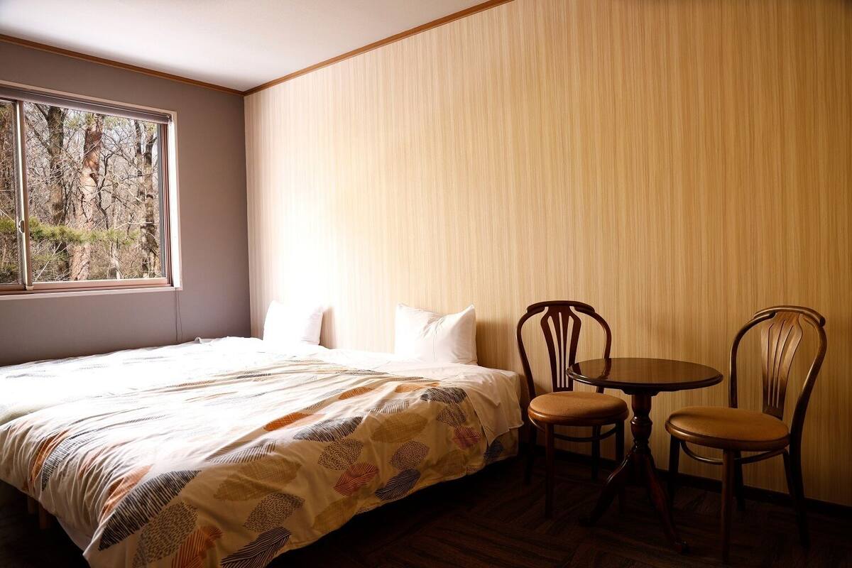 Un dormitorio con cama, dos sillas, una mesita y ventana con vista a árboles.