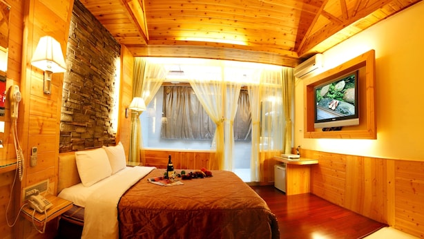 Una habitación acogedora de madera con una cama, un televisor, una mesita con una botella y vasos, y una ventana con cortinas.