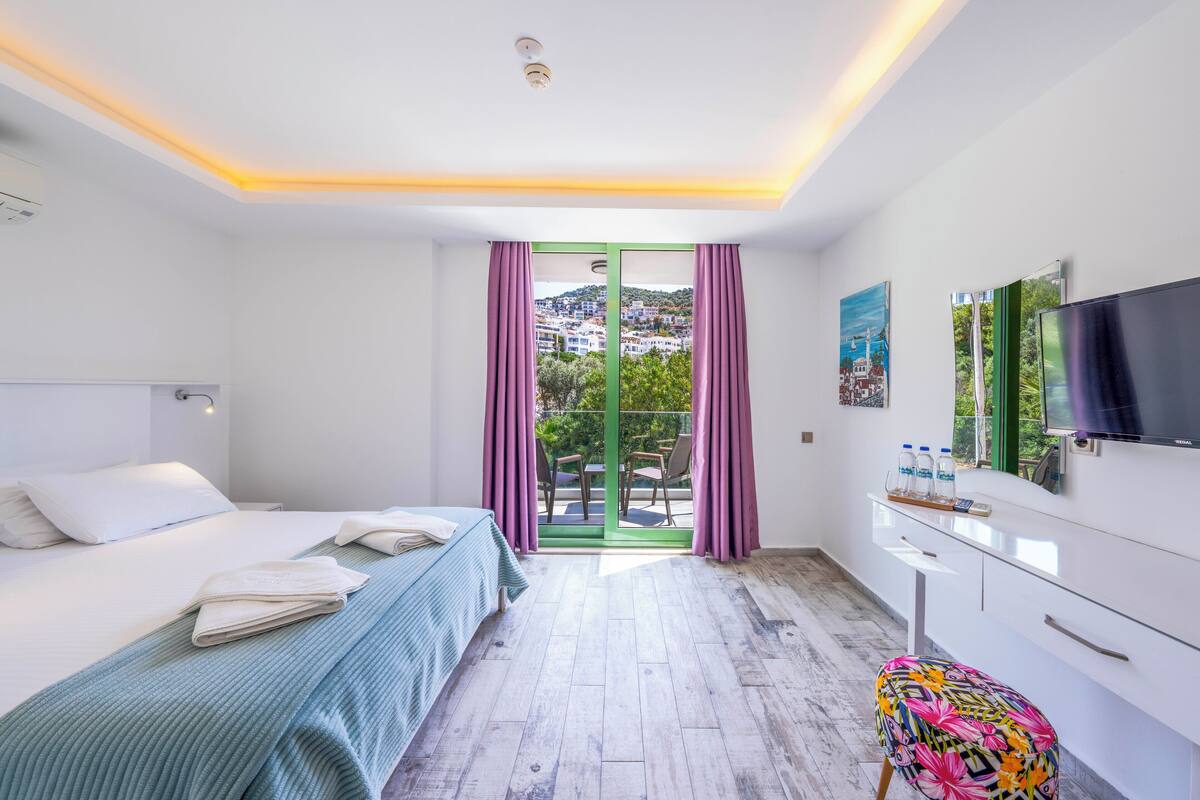 A modern hotel room with a bed, a desk, a TV, and a balcony with a view of the city.