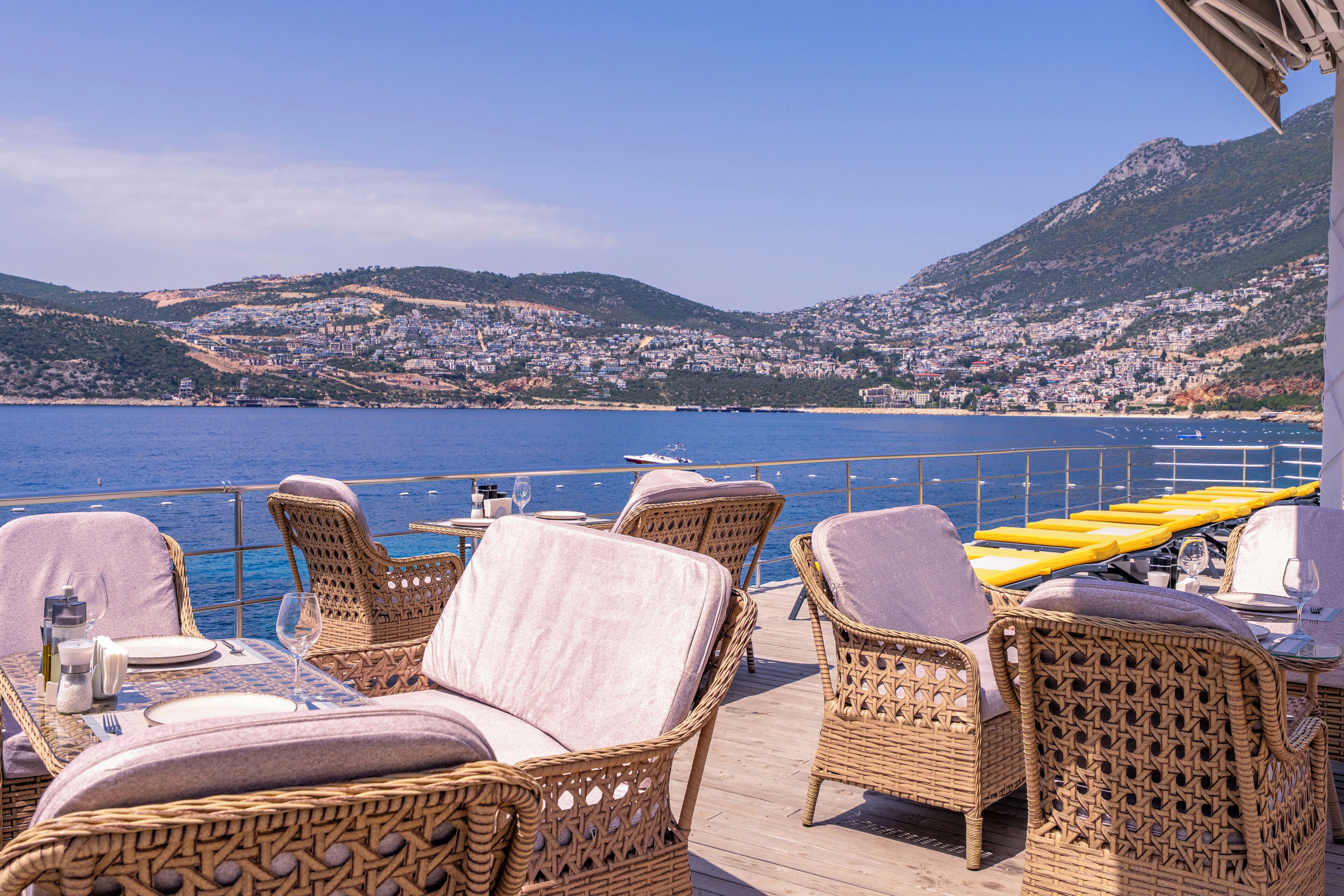 Una terraza con mobiliario de mimbre y una mesa lista para comer, con vistas a un pueblo costero y al mar.