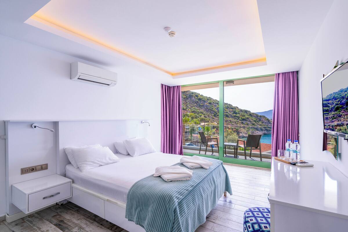 A modern hotel room with a large bed, a bedside table, a built-in air conditioning unit, a small desk with a mirror, and a balcony with a view of the hills and sea.