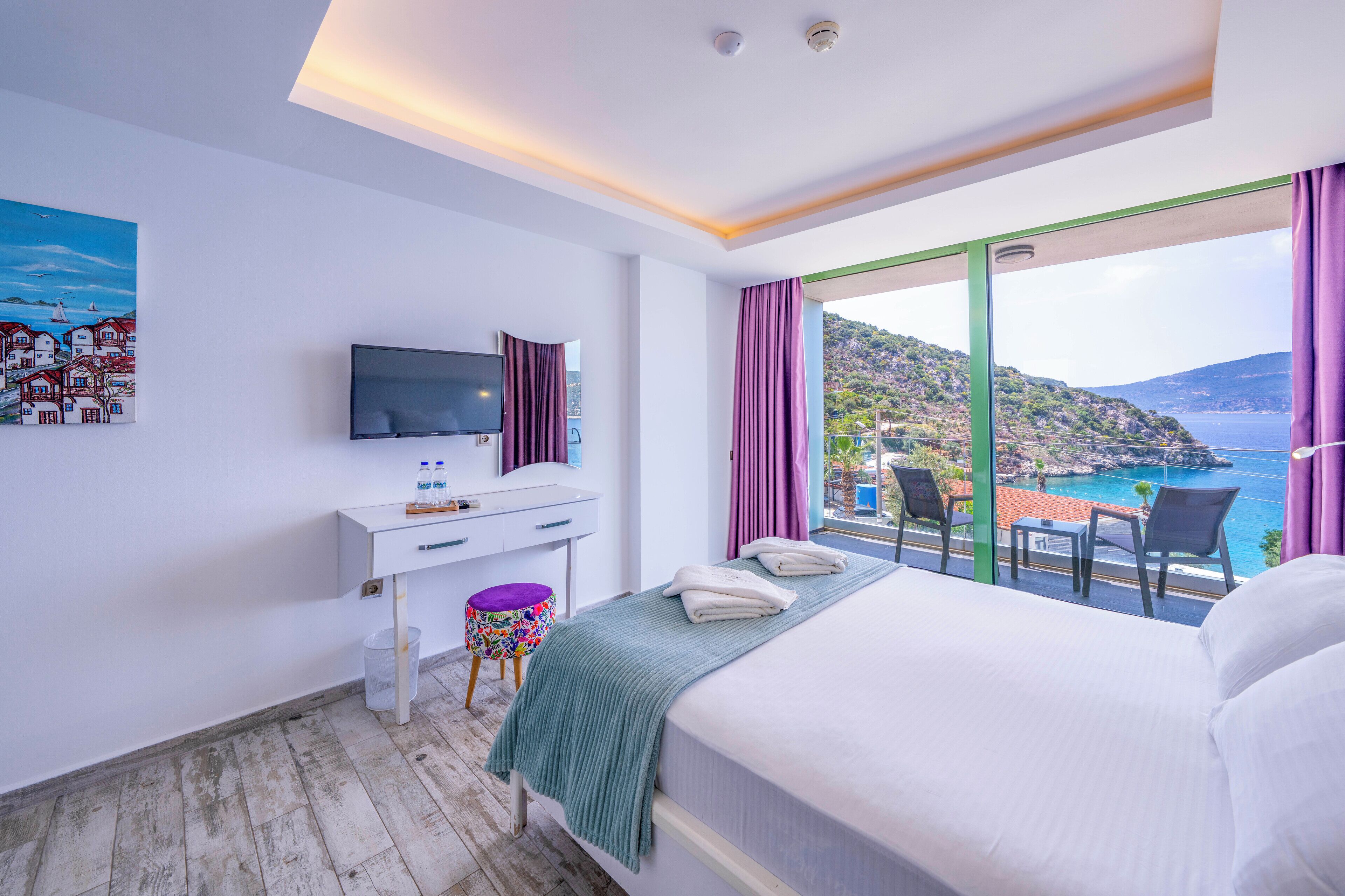 A hotel room with a bed, a desk with a TV, a small stool, and a view of the sea.