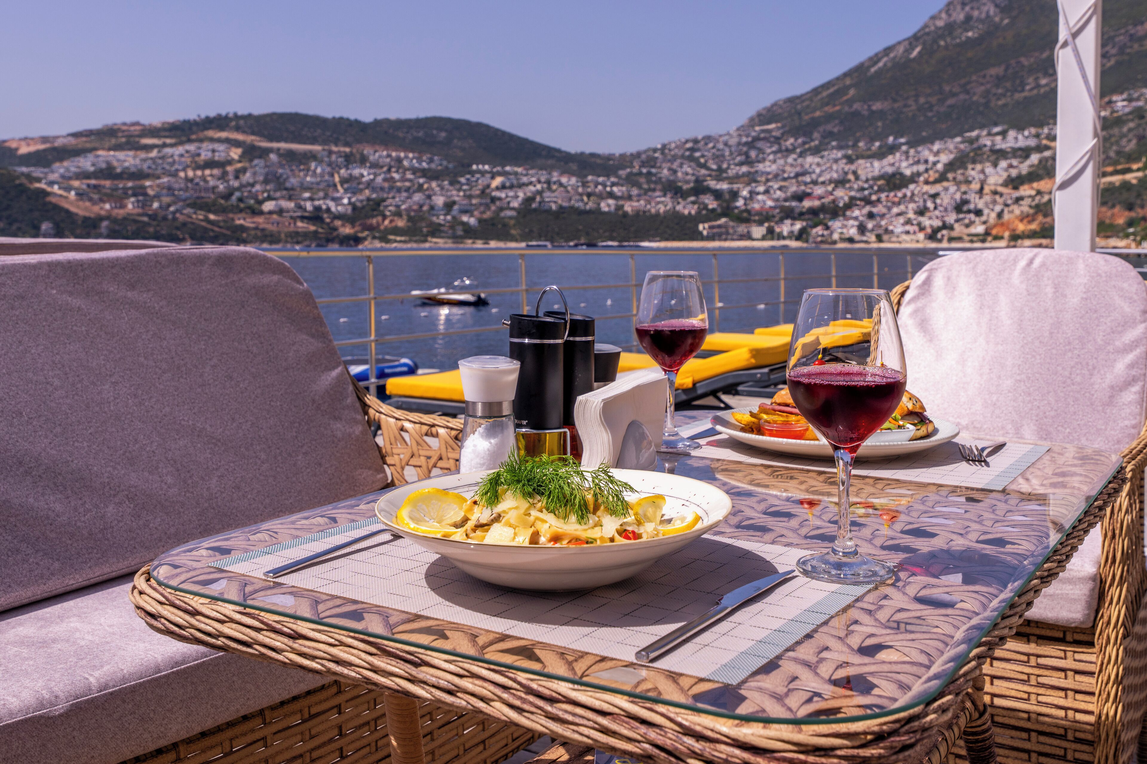 Una mesa de comedor con una silla de mimbre, un plato de comida y dos copas de vino tinto, con vistas a un pueblo costero.