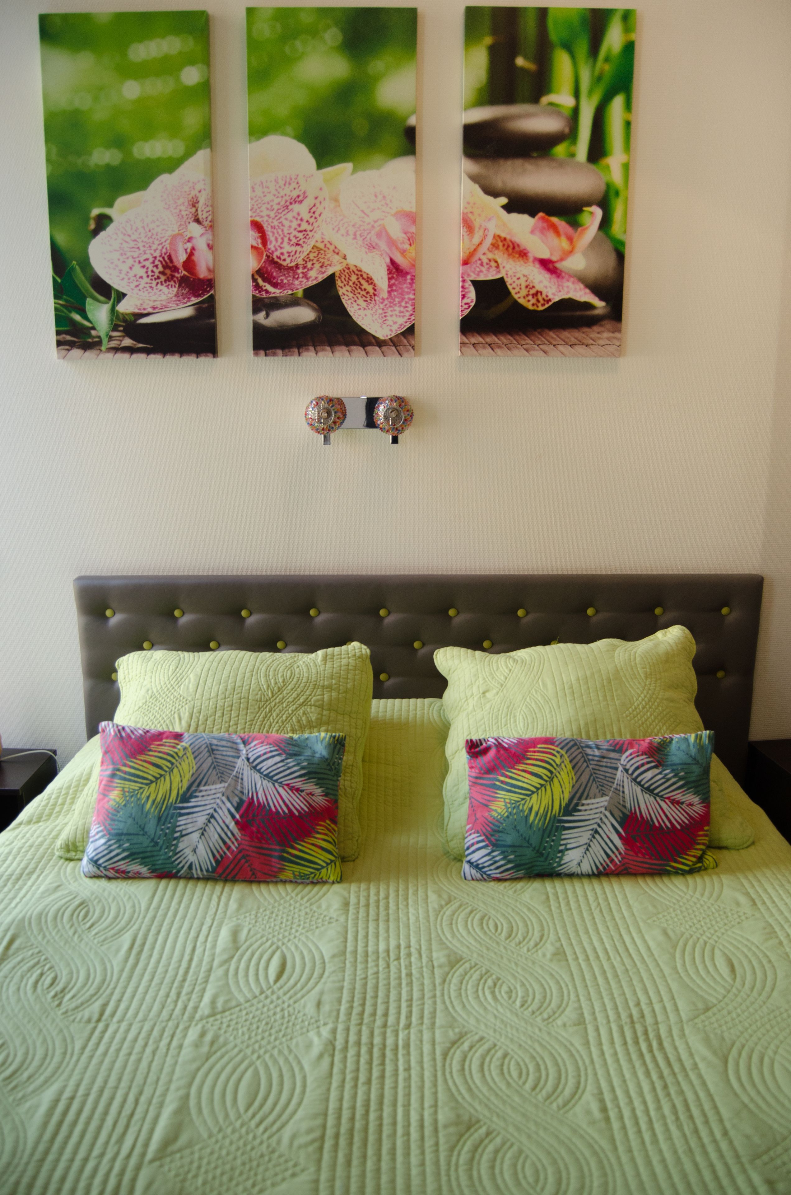 A bed with a green quilt and two pillows, a headboard, and three wall-mounted paintings of flowers and stones.