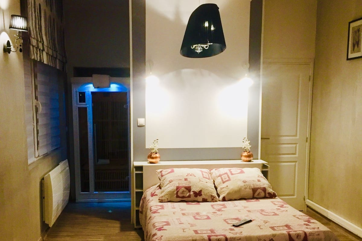 Ett sovrum med en s äng, sängbord, en hängande lampa och en dörr som leder till ett annat rum.