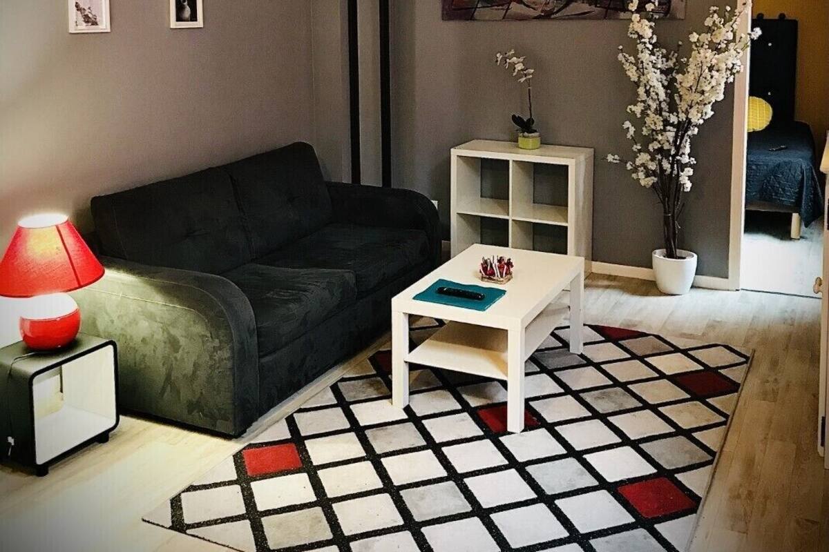 Ett vardagsrum med en svart soffa, ett vitt soffbord, en röd lampa och en mönstrad matta.