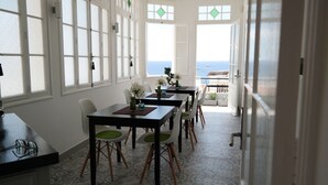 Un comedor con vista al mar, que presenta una mesa larga con sillas, paredes blancas y ventanas grandes.
