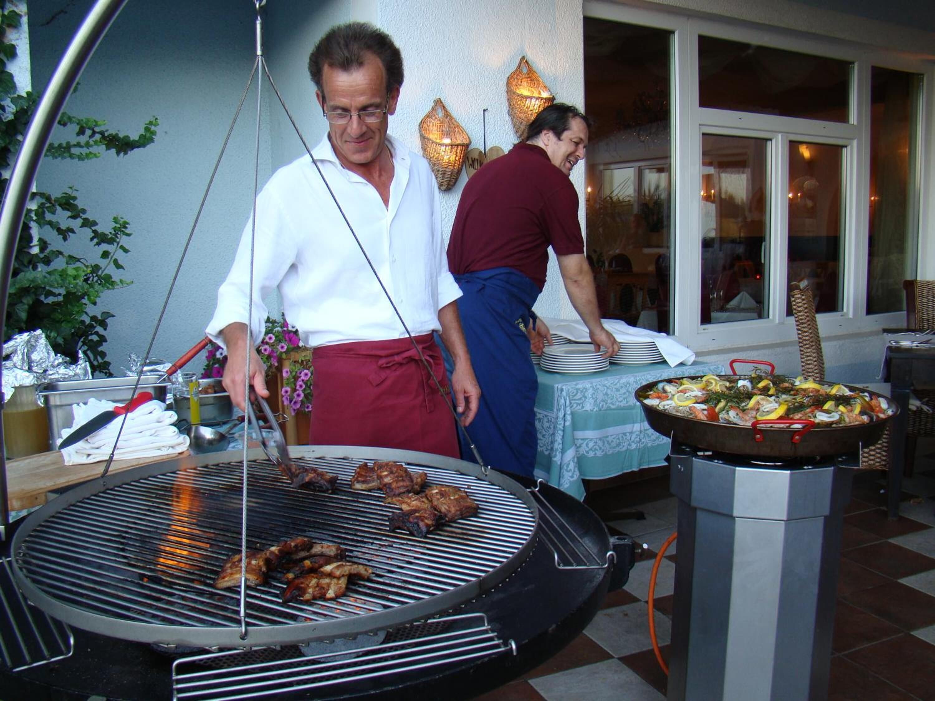 Un barbecue avec de la viande en train de cuire, une table avec des assiettes et un grand plat rempli de nourriture, et une personne portant un tablier rouge.
