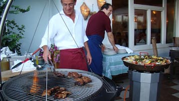 Un barbecue avec de la viande en train de cuire, une table avec des assiettes et un grand plat rempli de nourriture, et une personne portant un tablier rouge.