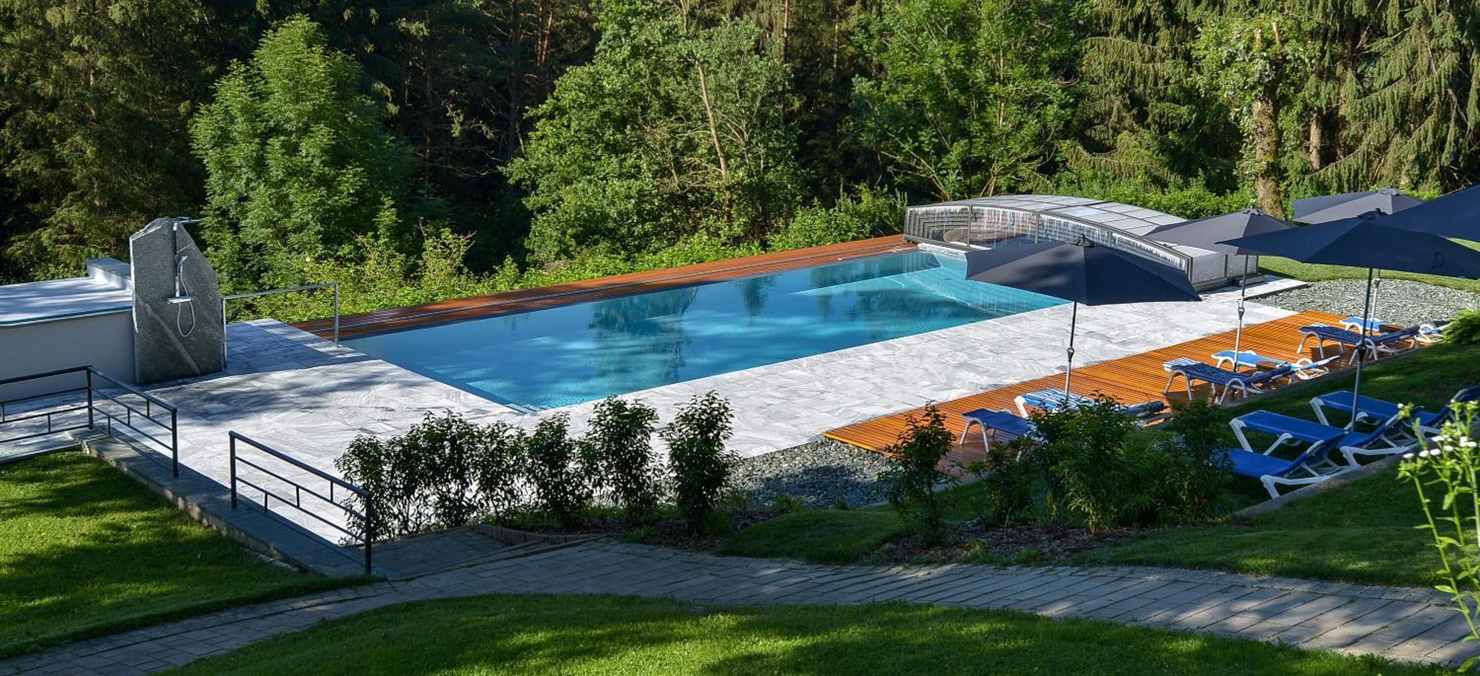 Ein Schwimmbecken mit Holzterrasse und Liegestühlen, umgeben von Grünpflanzen.