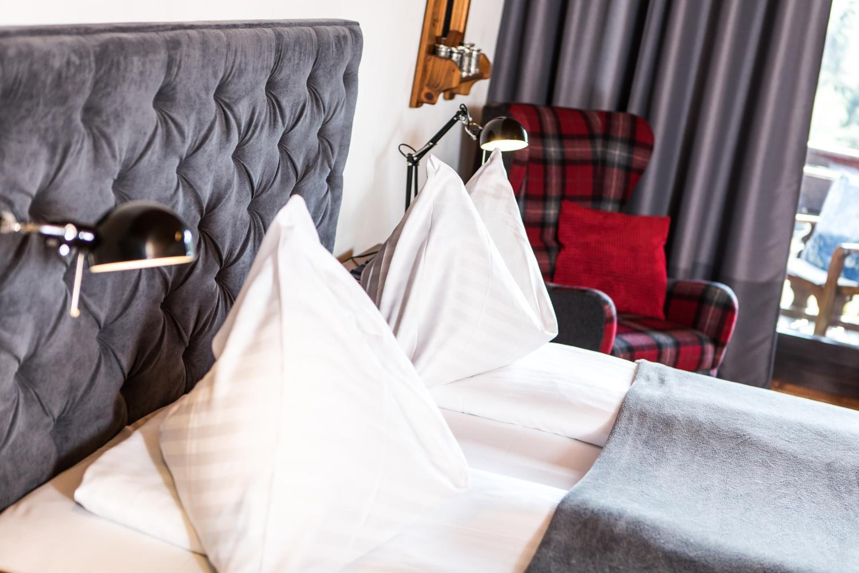 Una cama bien tendida con sábanas y almohadas blancas, un cabecero tapizado en gris y una silla de cuadros rojos al fondo.
