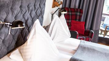 Una cama bien tendida con sábanas y almohadas blancas, un cabecero tapizado en gris y una silla de cuadros rojos al fondo.