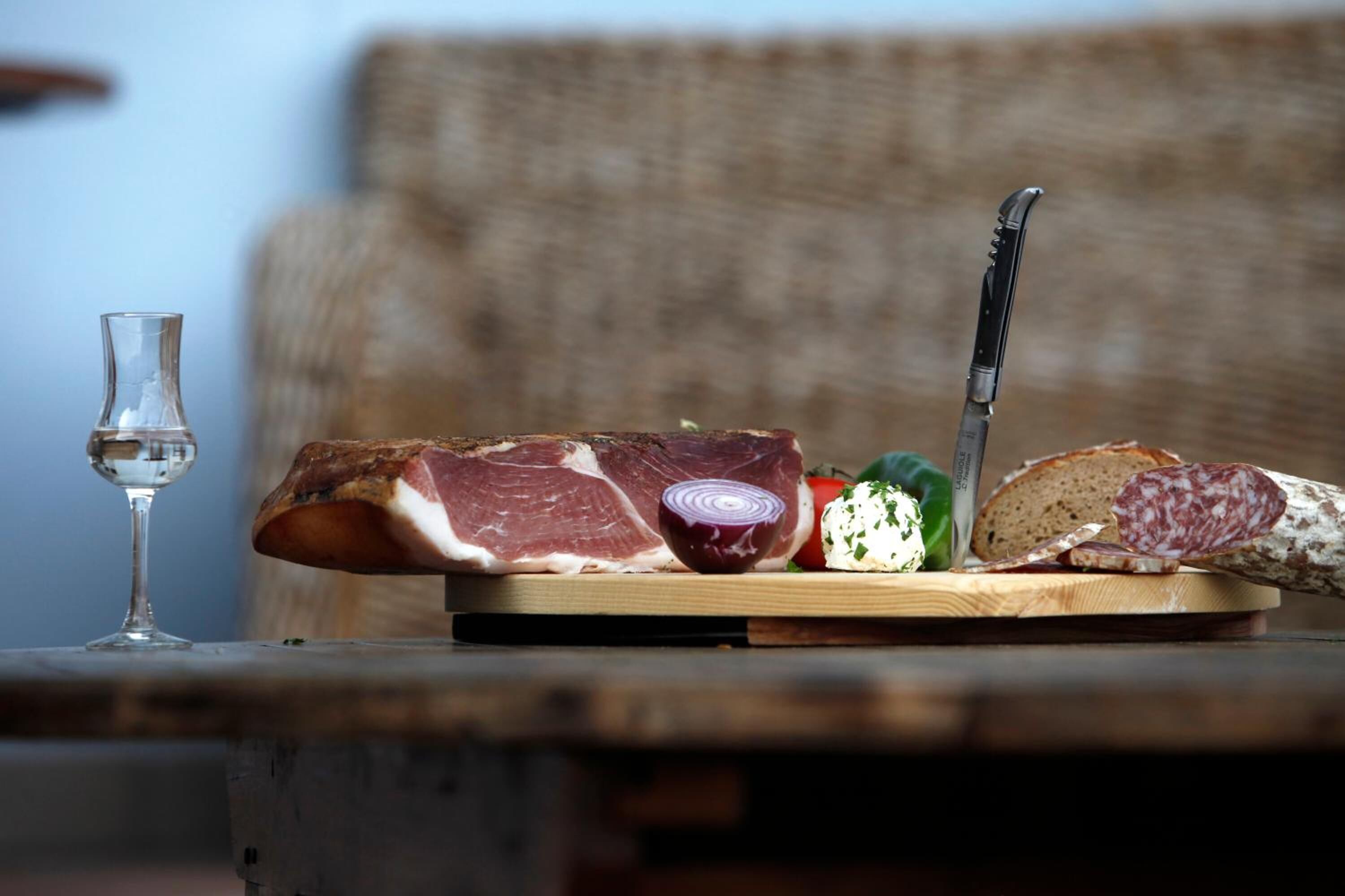 A wooden table with various food items including a loaf of bread, a piece of meat, a red onion, a green pepper, and sliced salami.