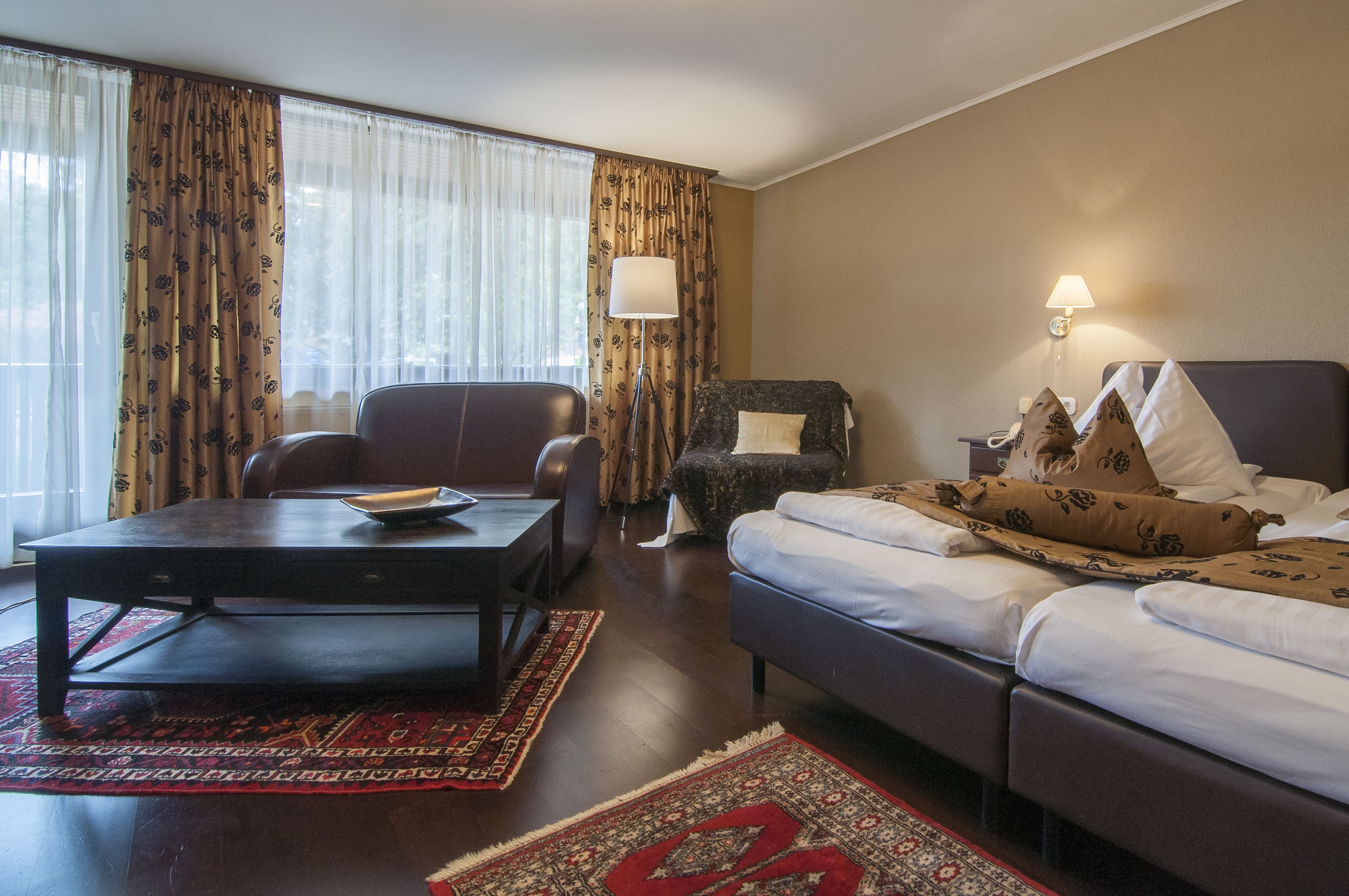 A hotel room with a bed, a sofa, a desk, and a chair. There is a lamp on the wall and a window with curtains.