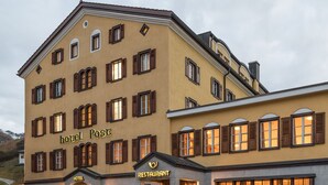 A multi-story building with a yellow facade, multiple windows with wooden shutters, and signage indicating it is a hotel and restaurant. - Hotel Post Bivio (Surses)