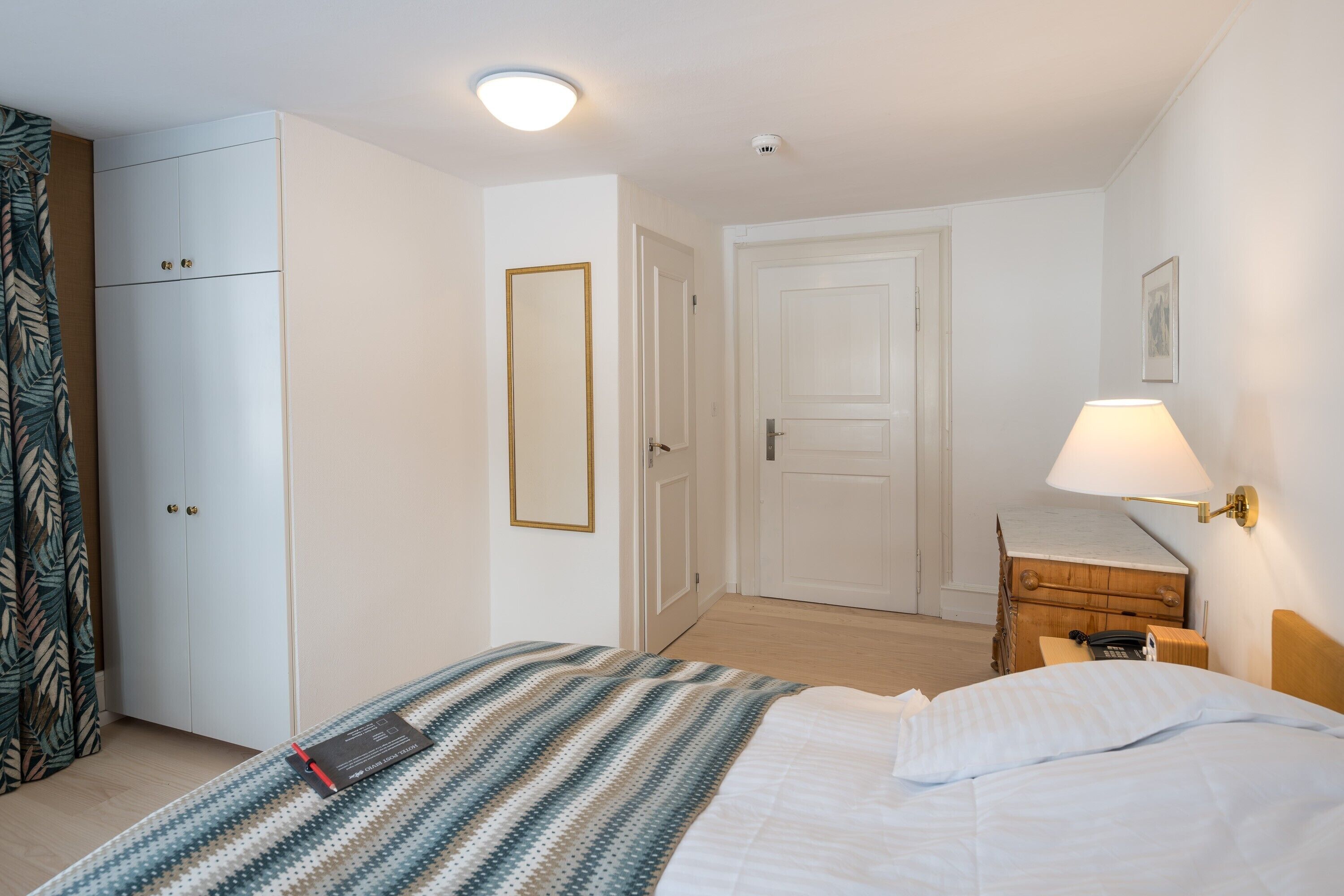 Una habitación de hotel con una cama, un armario, una mesita de noche con lámpara y un pequeño aparador de madera.
