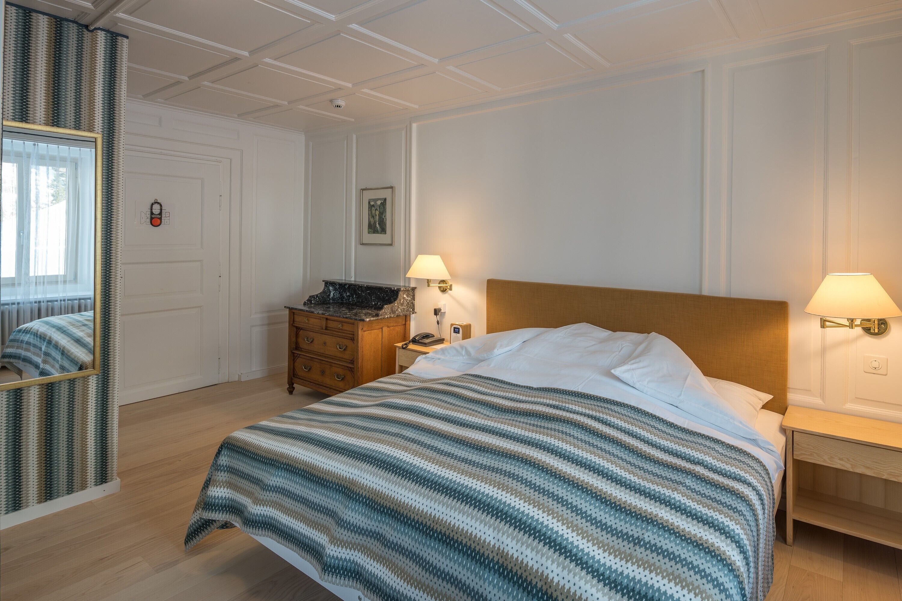 Une chambre d’hôtel avec un grand lit, deux tables de chevet avec des lampes, une commode en bois, un miroir et une porte équipée d’une caméra de surveillance.