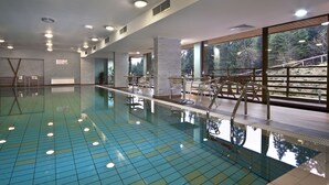 An indoor swimming pool with clear blue tiles, a tiled deck area, and large windows offering a view of trees and a wooden deck outside. - Club Hotel Yanakiev (Samokov)