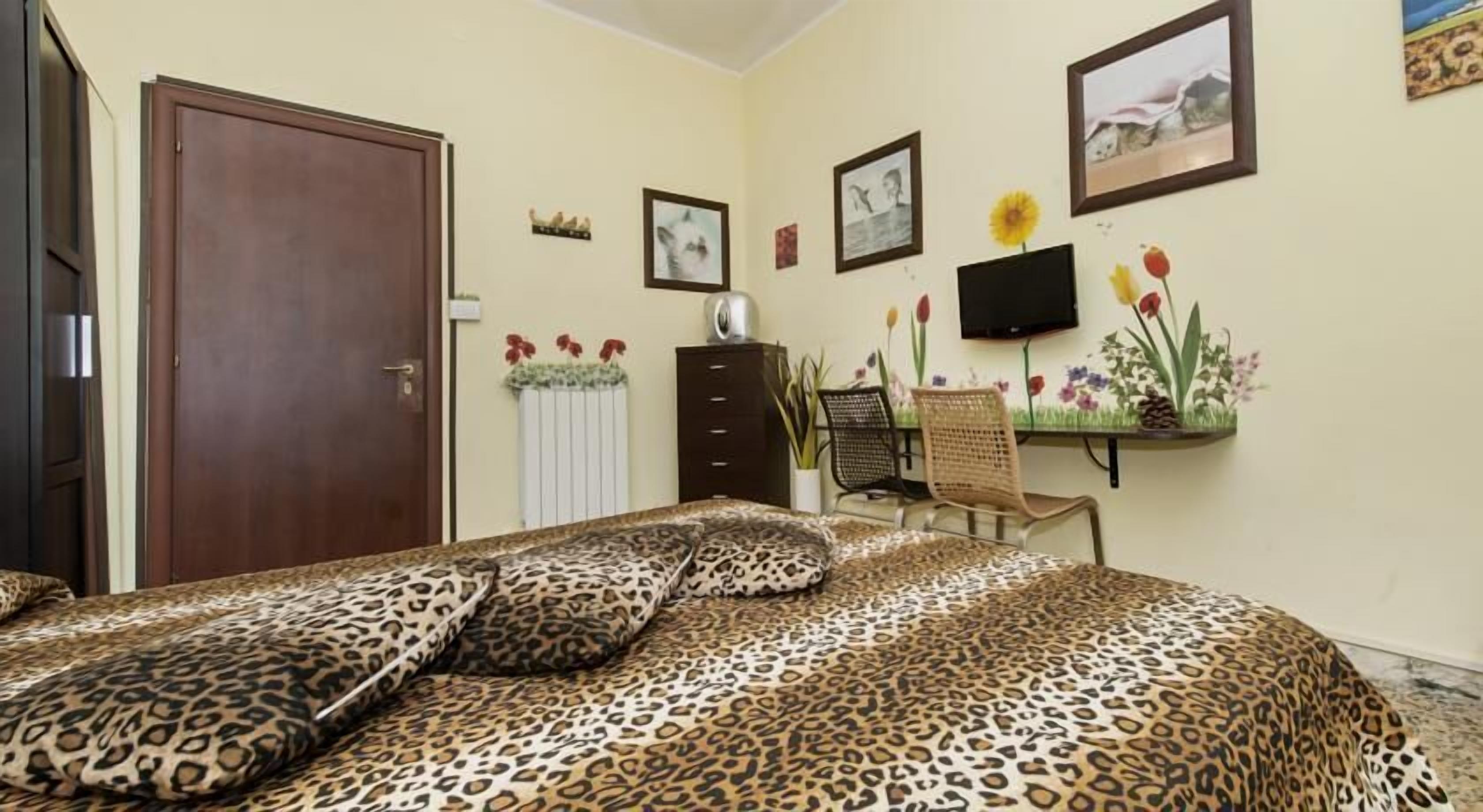 Une chambre avec une couvre-lit à motifs léopard, une porte en bois, une commode, une chaise, un téléviseur et des œuvres d’art murales.