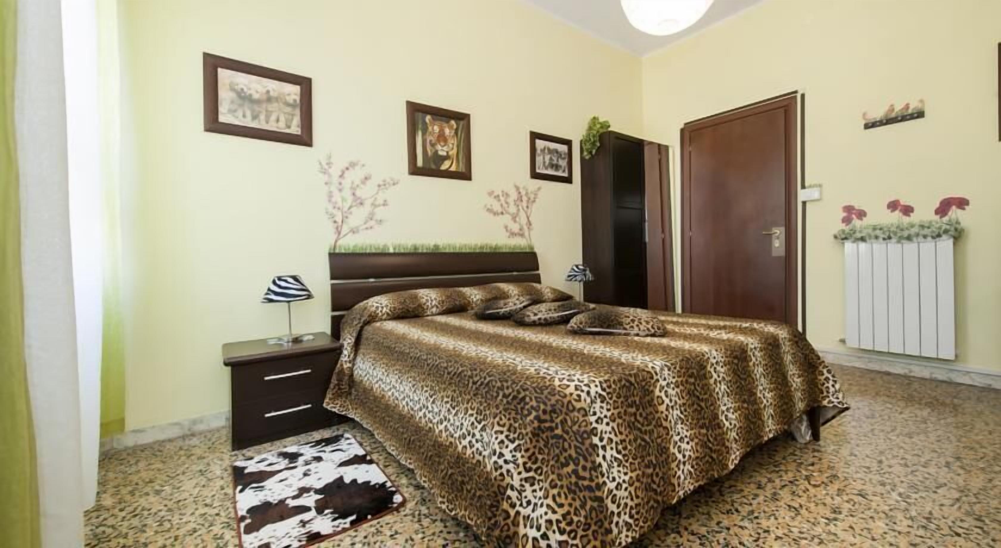 Une chambre avec une couvre-lit à motifs léopard, une table de chevet en bois avec une lampe, une porte et un radiateur décoré de fleurs.