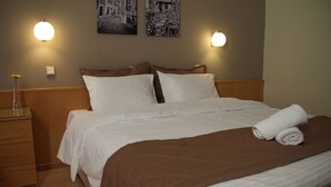Una cama bien hecha con ropa blanca y una manta marrón, una mesita de noche de madera con un jarrón de vidrio, y dos lámparas fijadas a la pared.