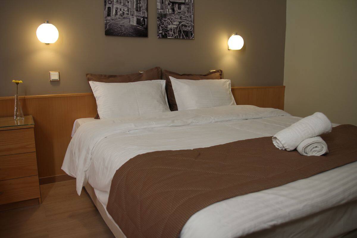 A neatly made bed with white linens and a brown throw blanket, a wooden nightstand with a glass vase, and two wall-mounted light fixtures.