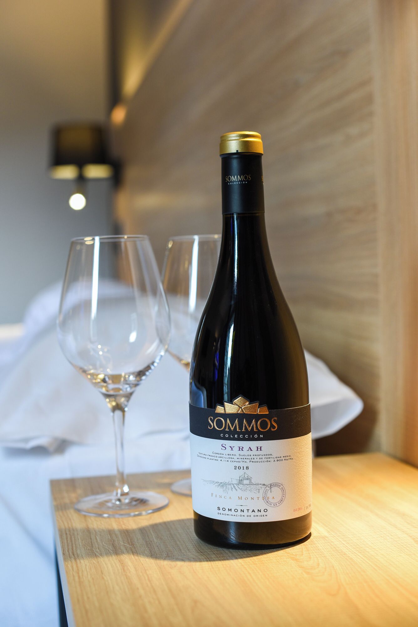 A bottle of SOMMOS Syrah wine with a label, placed on a wooden surface with two empty wine glasses beside it.