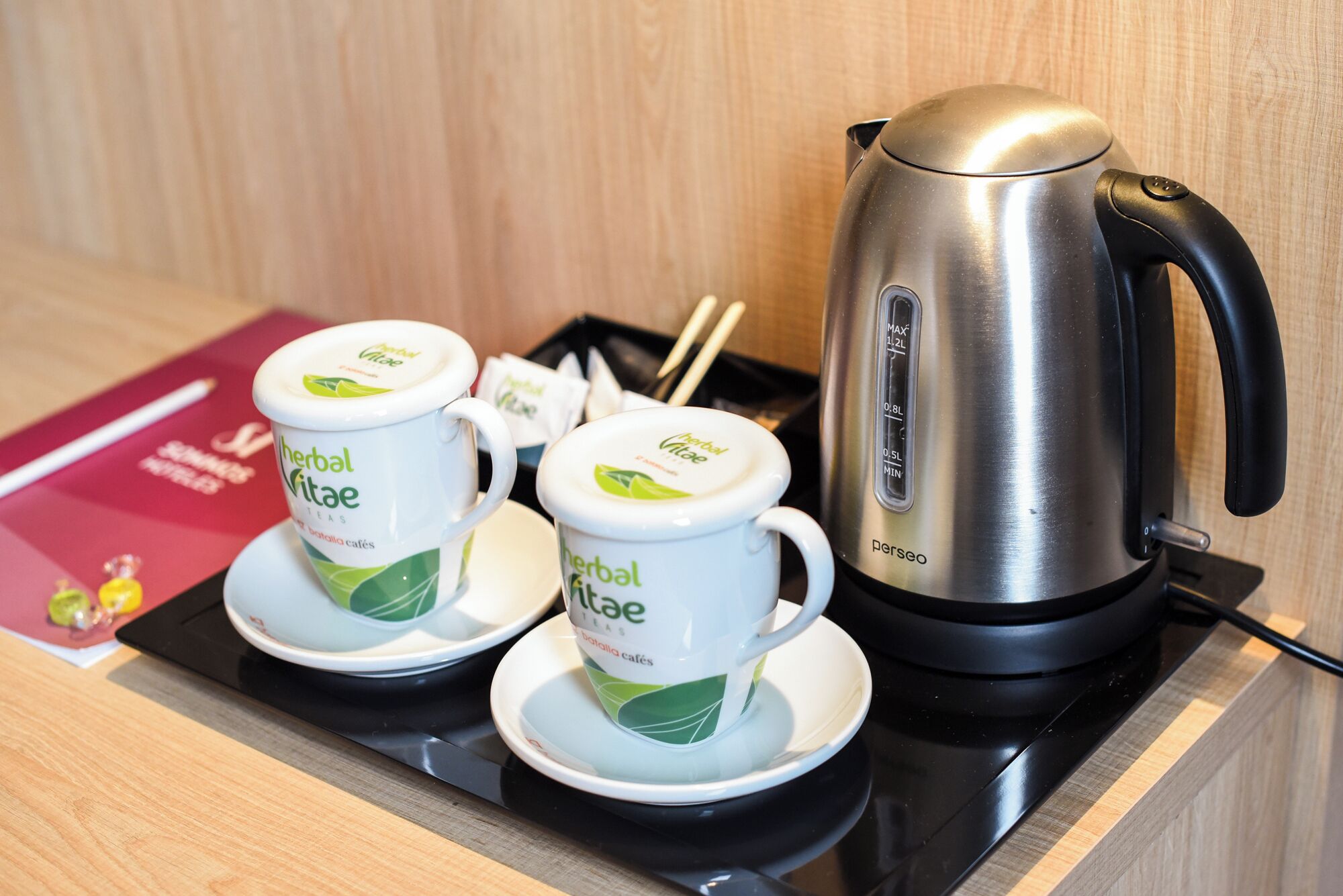 A tea station with a stainless steel electric kettle and three white cups with green leaf designs, labeled "herbal vitality teas."