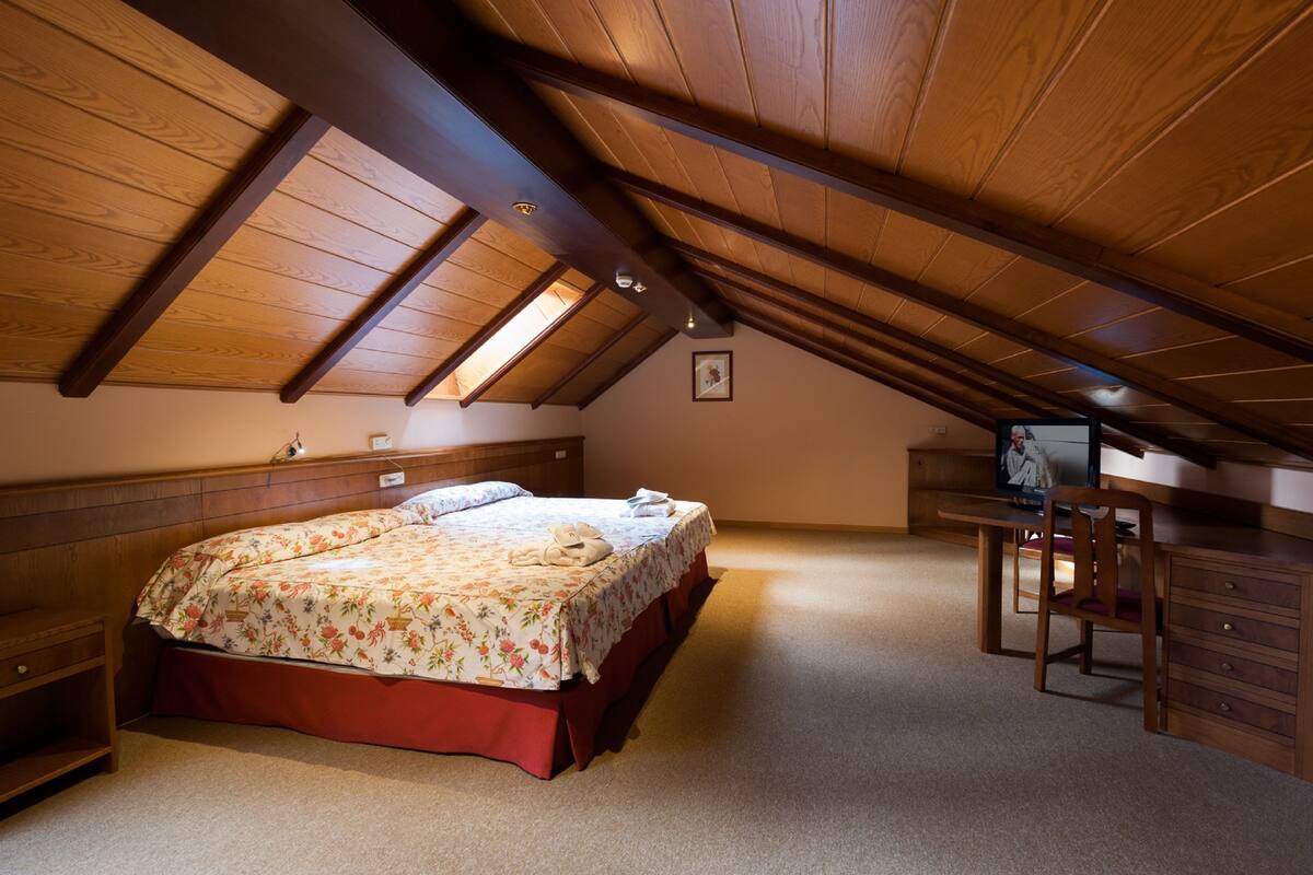 Un dormitorio con techo inclinado, una cama con ropa de cama floral, un escritorio con una silla y un televisor.